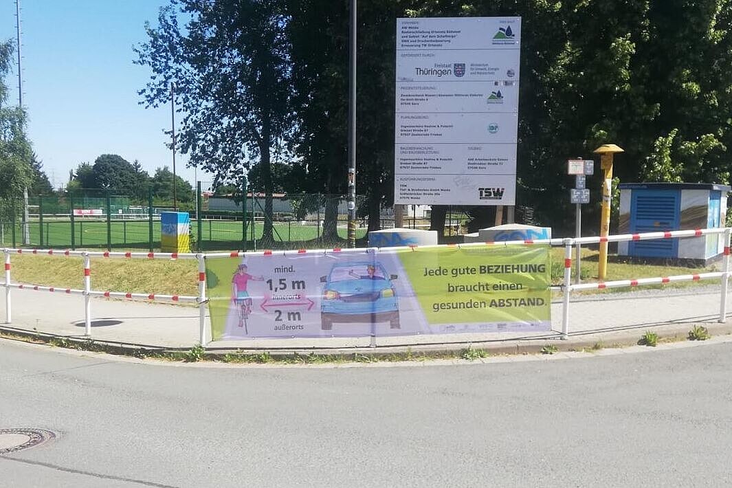 Ein Abstandsbanner hängt an einem Zaun an der Straße.